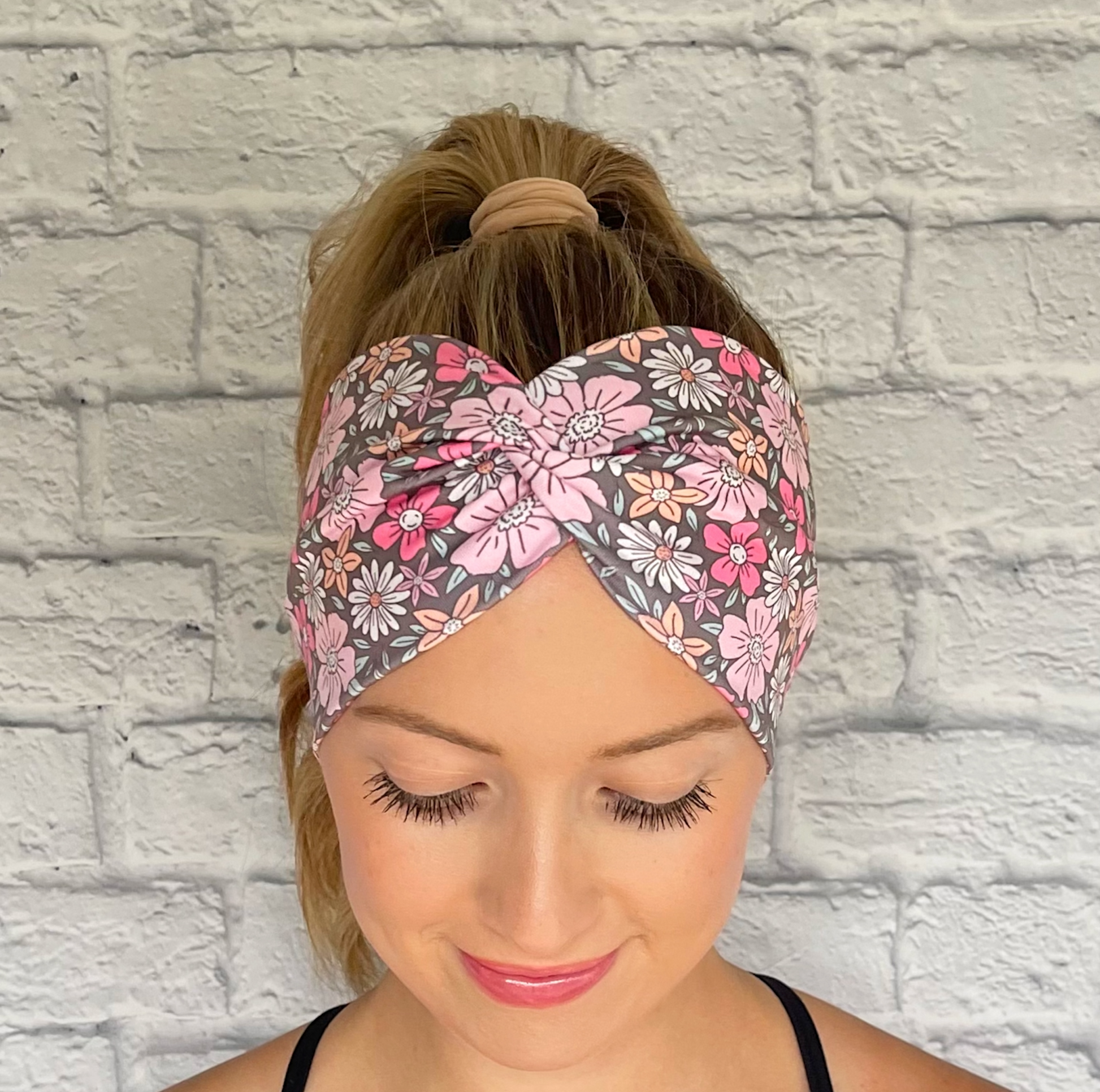 Woman with hair in curled ponytail wearing wide, twisted headband in gray with pink, peach, and white floral print.