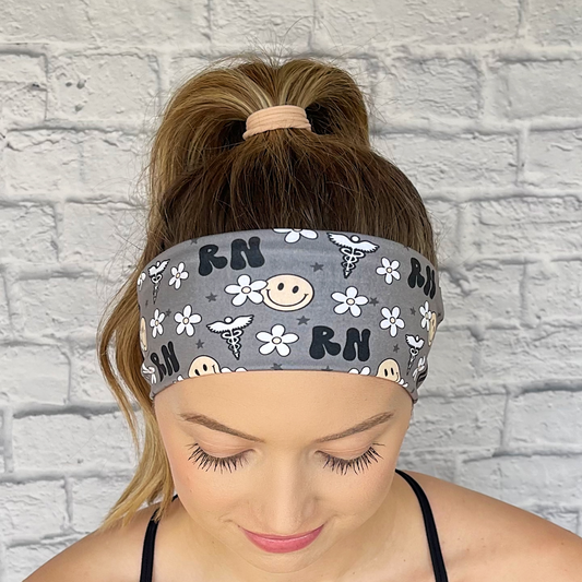 Woman with hair in curled ponytail wearing gray, stretchy headband with black "RN" print, yellow smiley faces, white flowers, and white medical theme print.