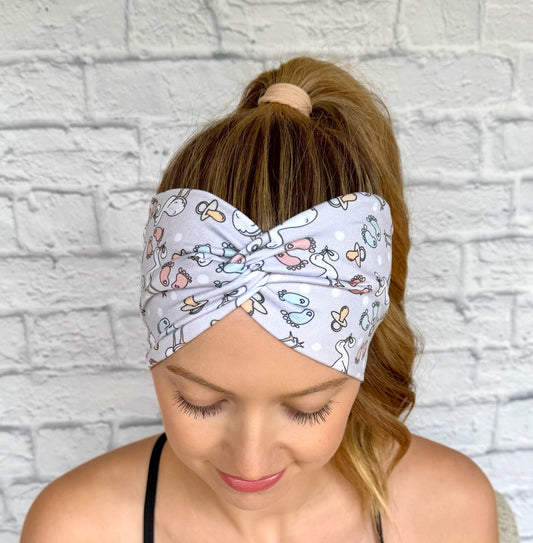 Woman with hair in curled ponytail wearing wide, twisted, light gray headband with stork and baby theme print.