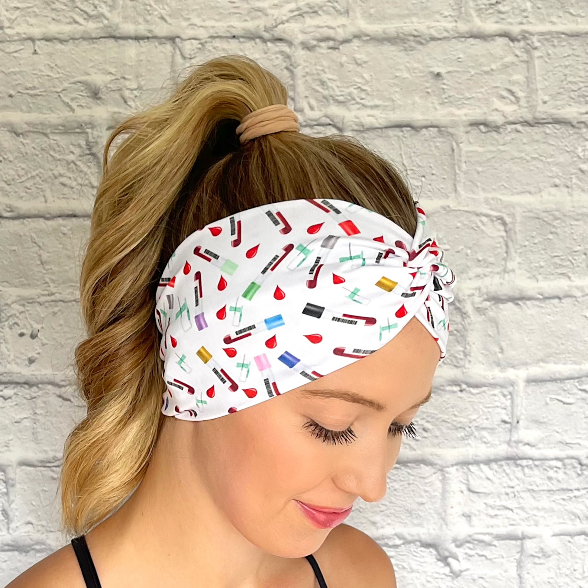 Woman with hair in ponytail wearing white, wide twist headband with phlebotomy theme print.