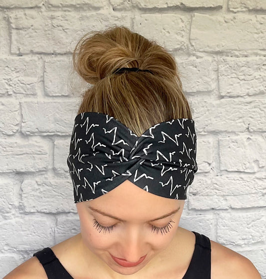 Woman with hair in bun wearing black, wide, stretchy, twisted headband with white EKG print.