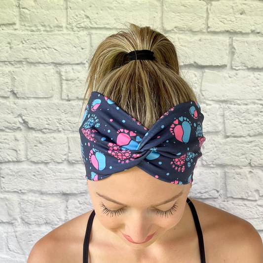 Woman wearing navy blue, wide twist headband with pink and blue baby feet.