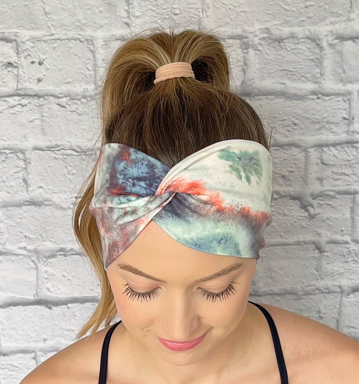 Woman with hair in curled ponytail wearing wide, twisted headband in tie dye print with peach, white, light blue, and dark gray/blue.