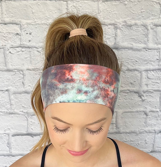 Woman with hair in curled ponytail wearing flat, stretchy headband in pink, white, blue, and gray/purple tie dye print.