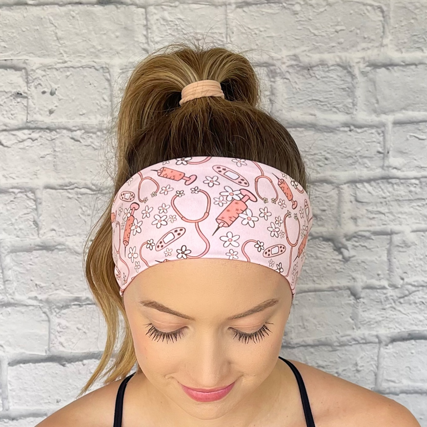 Woman with hair in curled ponytail wearing pink headband with pink and white medical theme print- stethoscopes, bandages, syringes, and flowers.