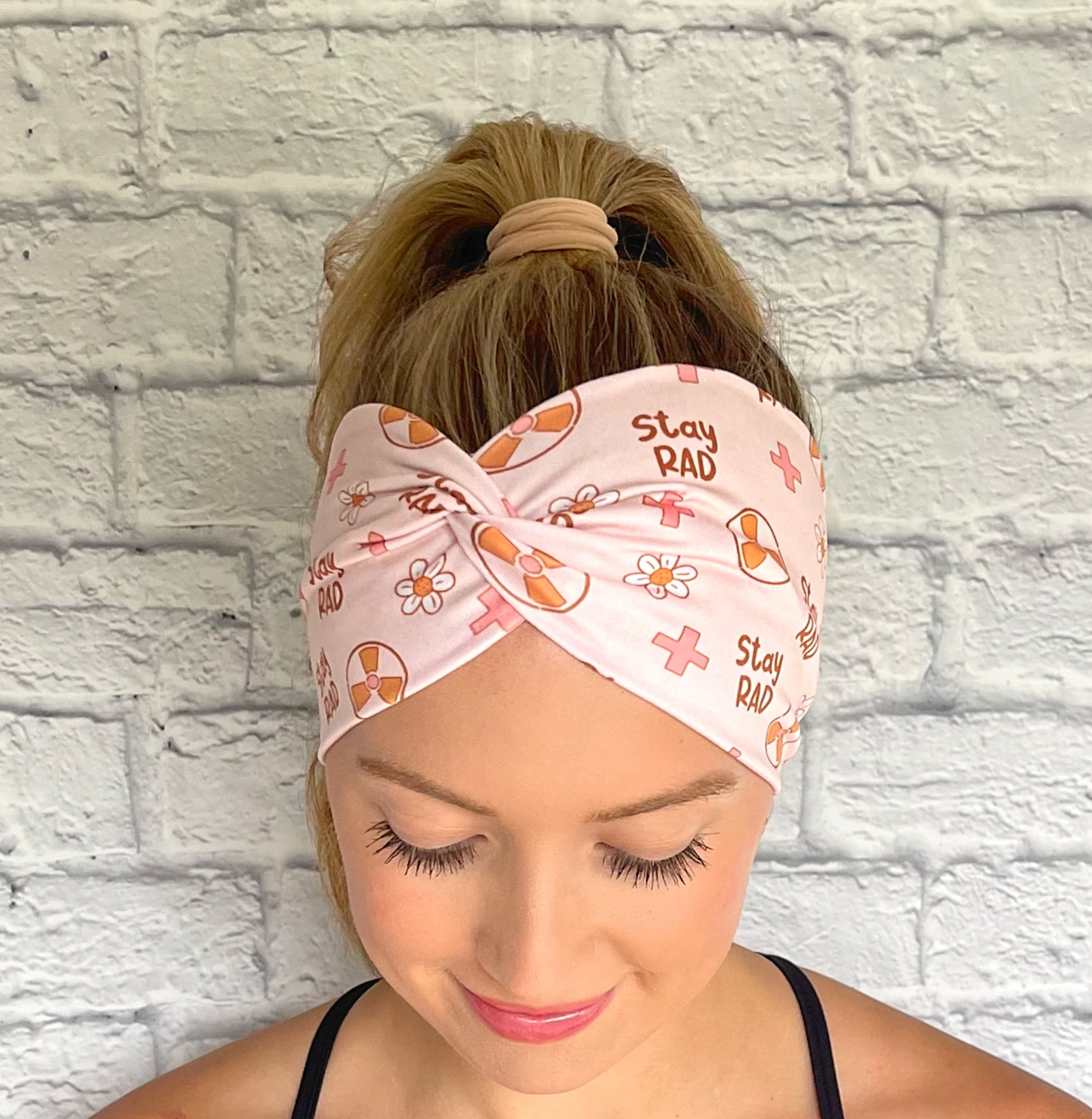 Woman with hair in curled ponytail wearing, twisted, wide headband in pink with "stay rad" print, radiology theme print, and flowers.