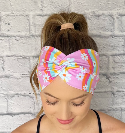 Woman with hair in curled ponytail wearing wide, twisted headband in light pink with white flower and swirl print in light blue, yellow, dark pink, and orange.