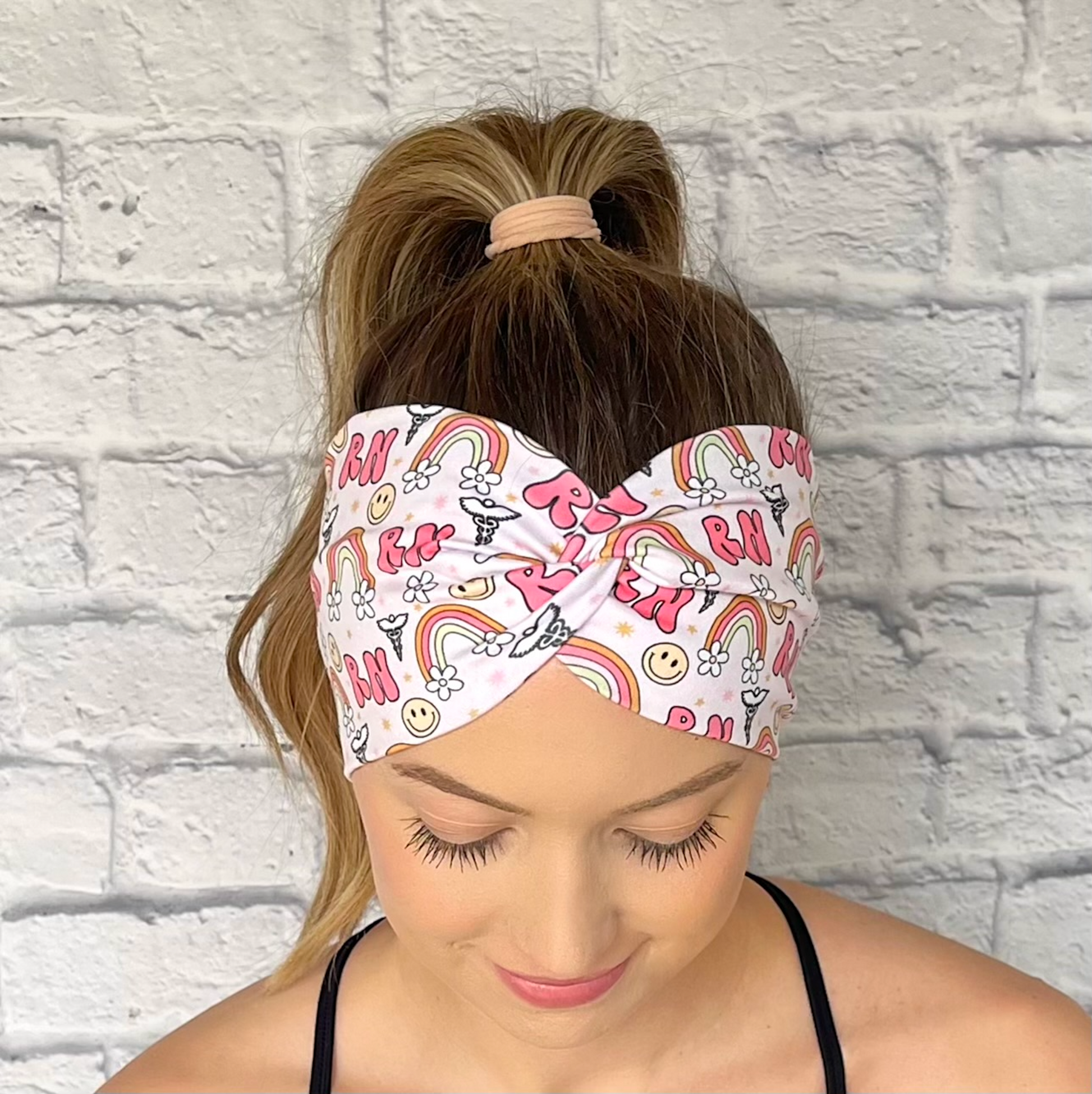 Woman with hair in curled ponytail wearing wide, twisted headband with pink "RN" print, rainbows, flowers, smiley faces, and medical symbol.