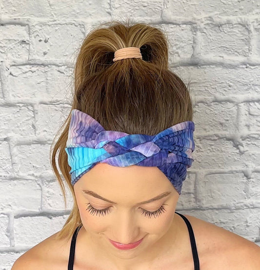Woman with hair in curled ponytail wearing sailor knot style headband in ribbed fabric with purple and blue tie dye print.