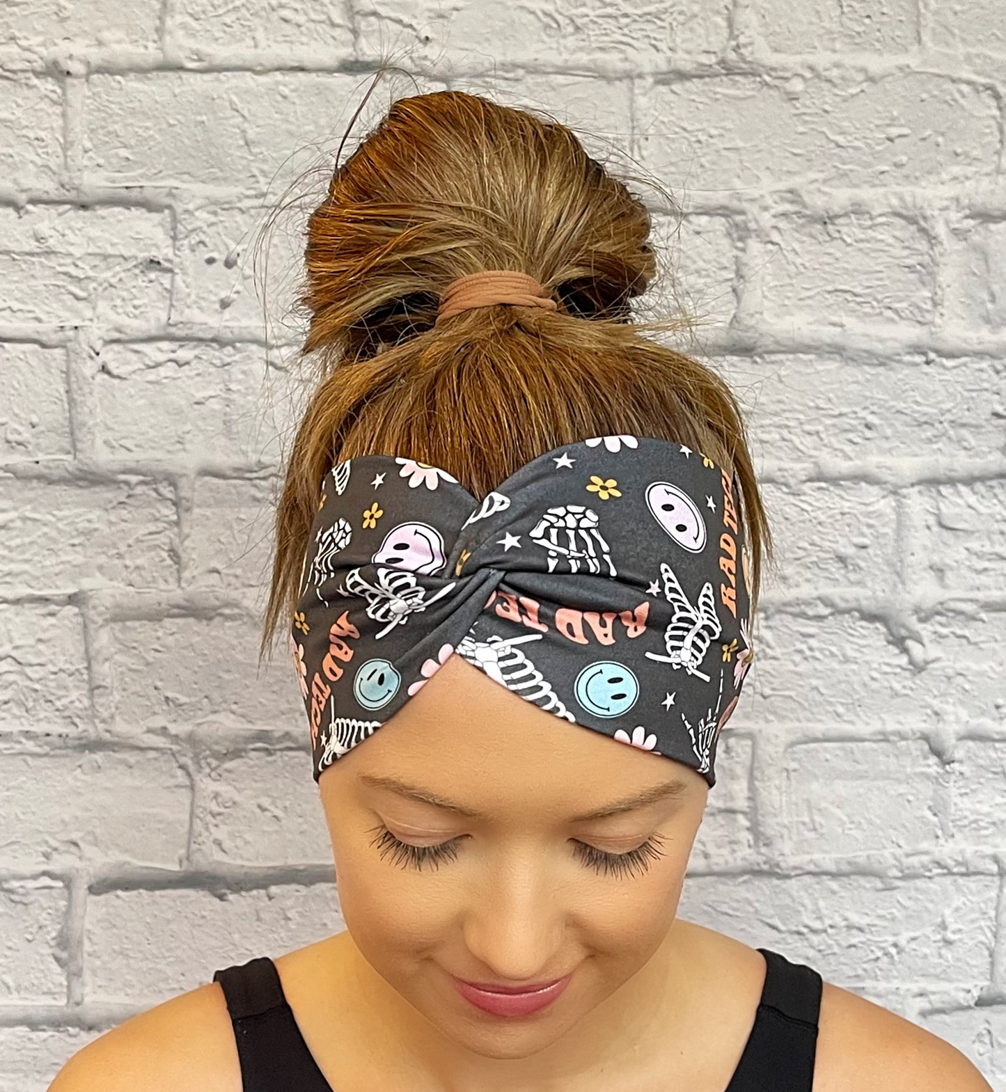 Woman with hair in bun wearing a dark gray, wide, twisted headband with skeletons, flowers, smiley faces, and "rad tech" print.