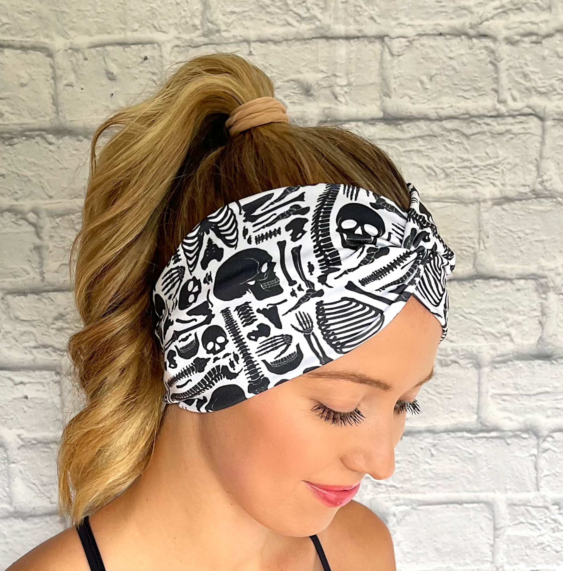 Woman with hair in curled ponytail wearing white, wide twist headband with black skeleton print. 