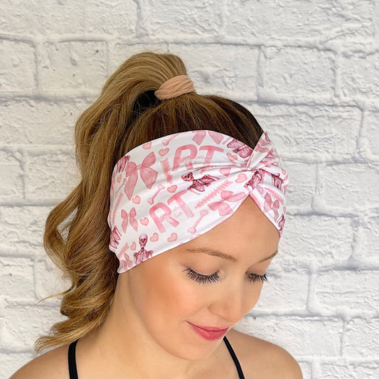 Woman with hair in curled ponytail wearing wide, white, twisted headband with pink "RT" and "radiology tech" print, pink bows, pink hearts, and pink skeletons.