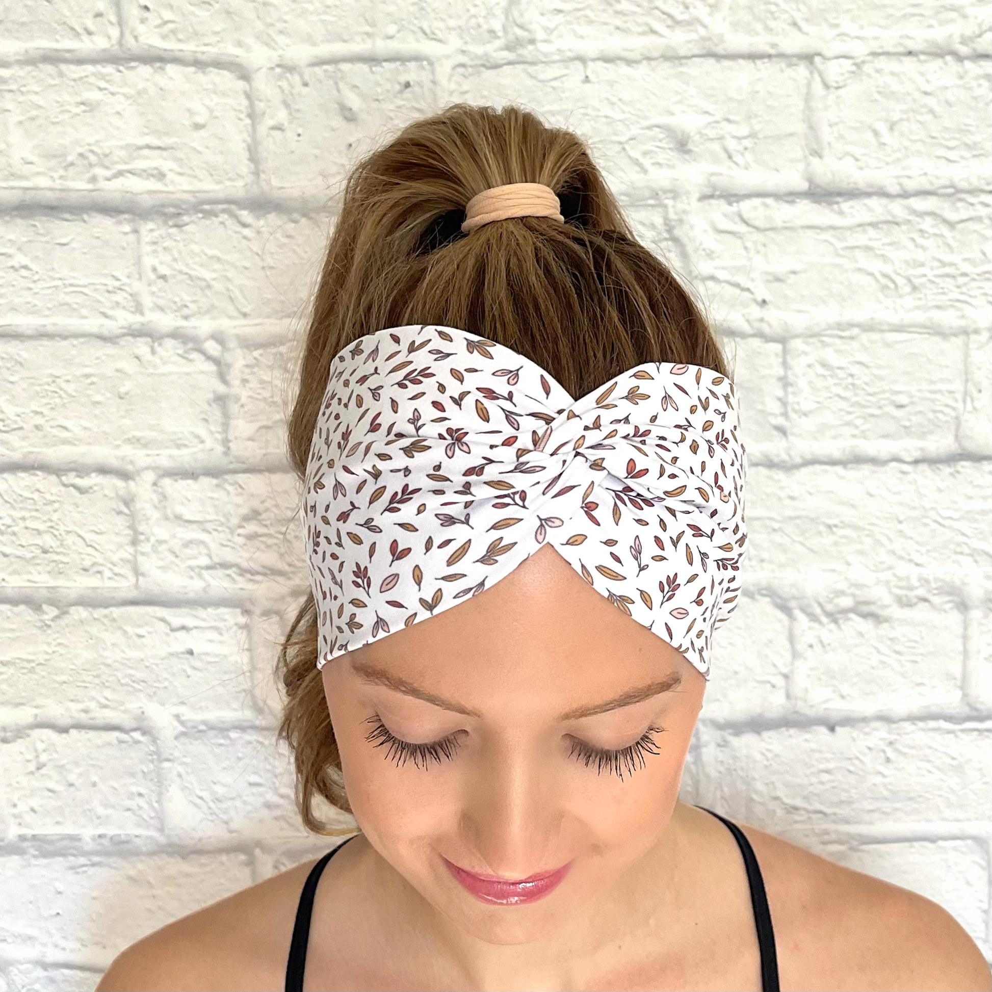 Woman with hair in ponytail wearing white, wide, twisted headband with brown/orange small leaves print.