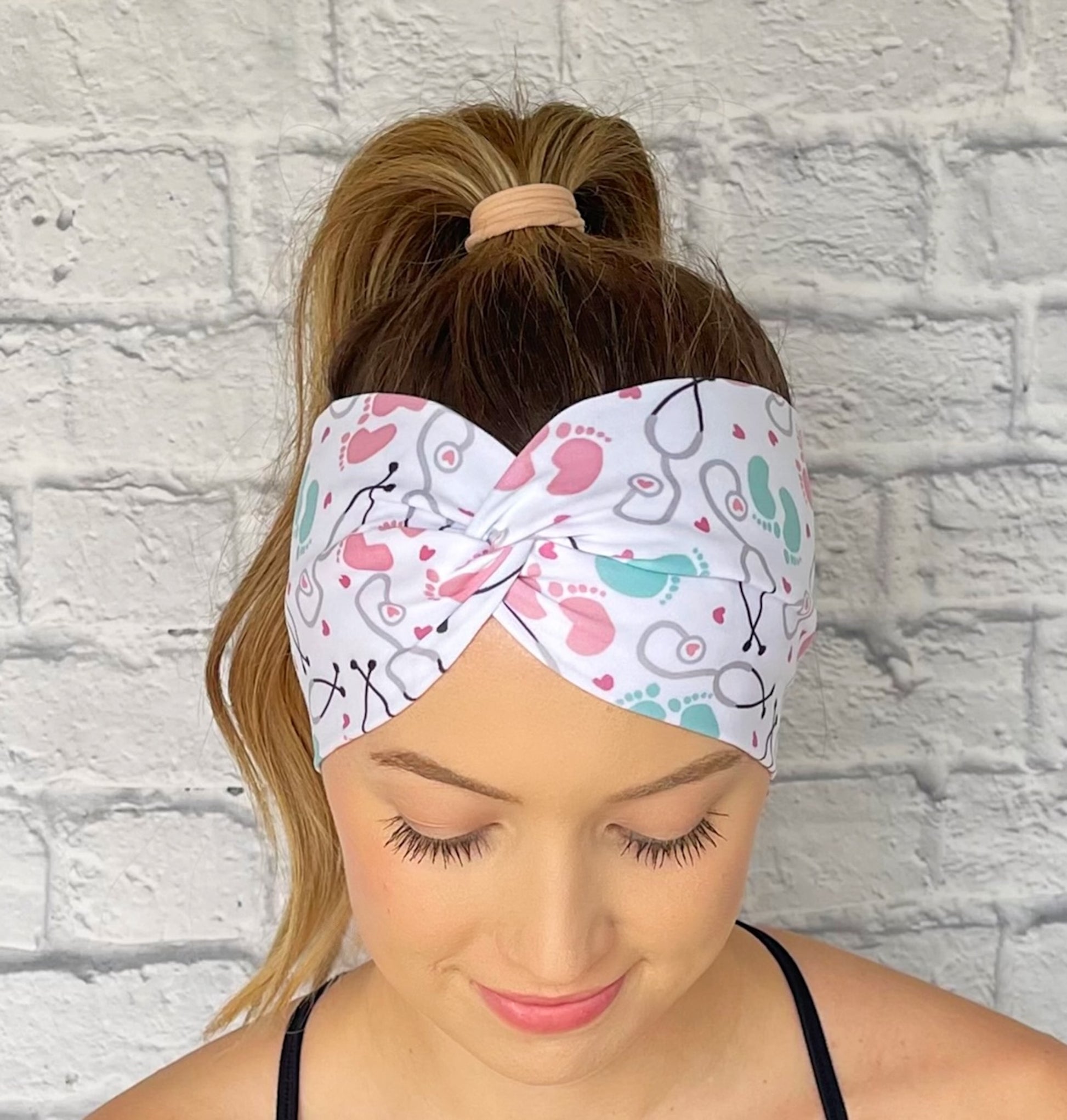 Woman wearing a white, OBGYN baby themed headband against a white brick wall.