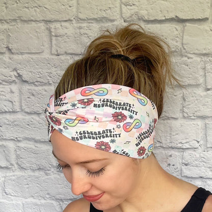 Woman with hair in bun wearing wide, twisted headband in rainbow plaid print with "celebrate neurodiversity", rainbow infinity signs, flowers, and lightning bolt print.