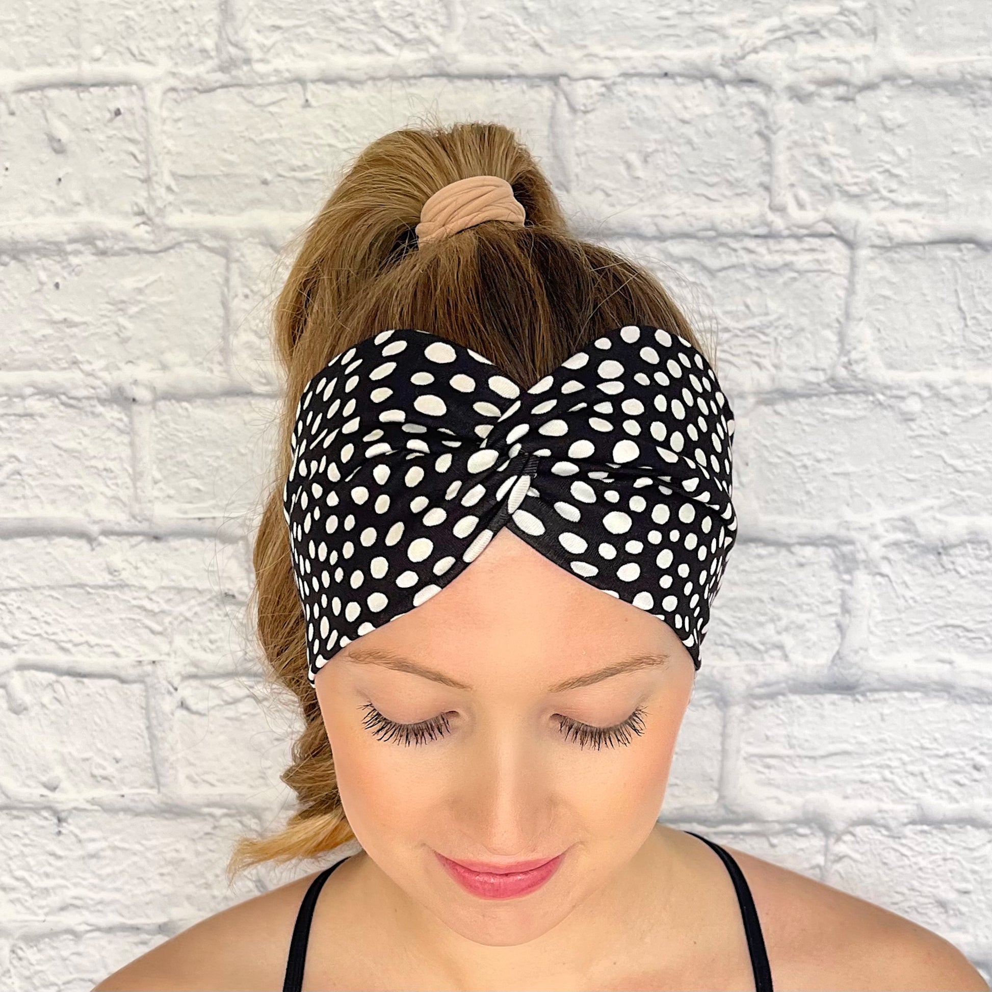 Woman with hair in curled ponytail wearing wide, twist headband in black with white polka dot print.