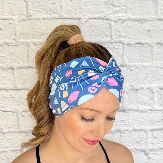 Woman with hair in curled ponytail wearing wide, twisted, blue headband with "OT" print and occupational therapy accessories theme print.