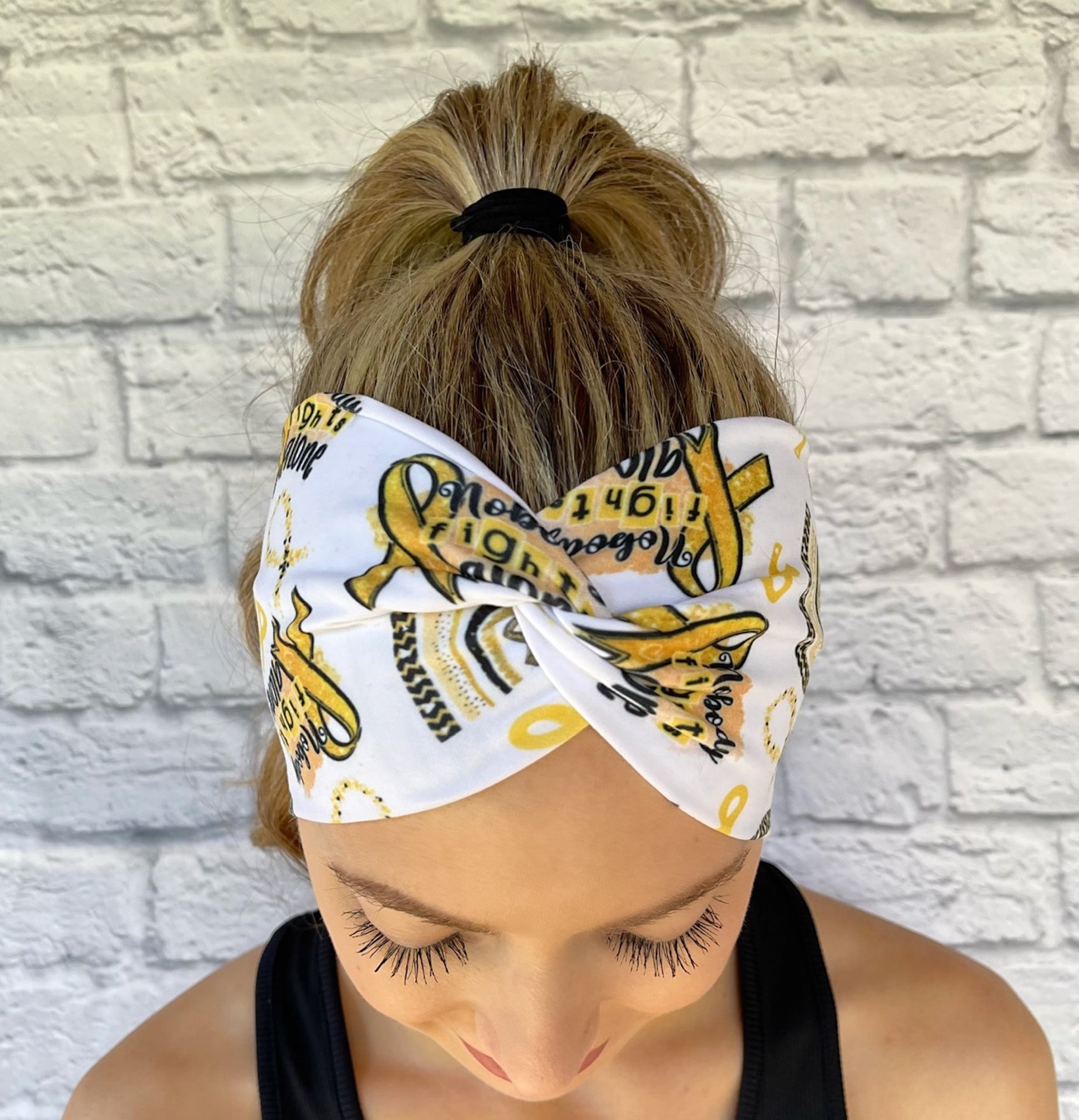 Woman with hair in ponytail wearing wide, twisted, white headband with "no one fights alone", gold ribbons, and gold/black rainbow print.