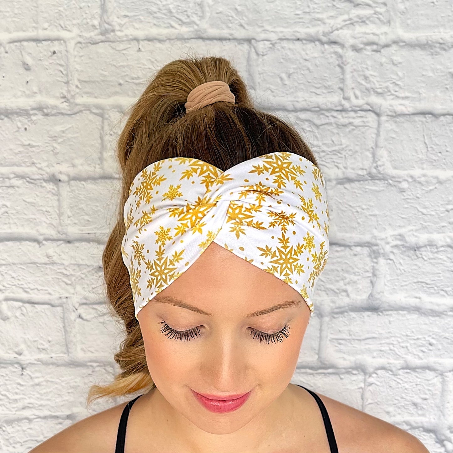 Woman with hair in curled ponytail wearing wide, white, twisted headband with gold color snowflake print.