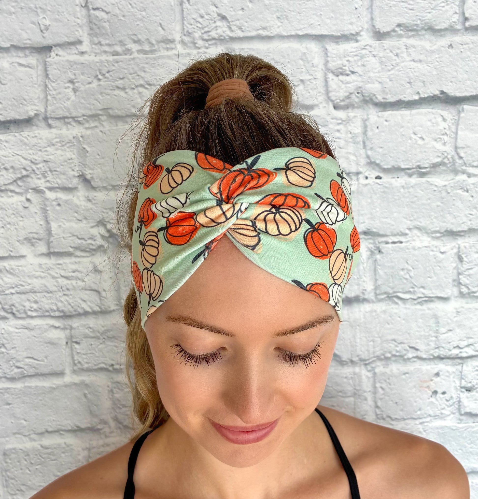 Woman with hair in ponytail wearing wide, twisted, stretchy, mint green headband with pumpkin print.