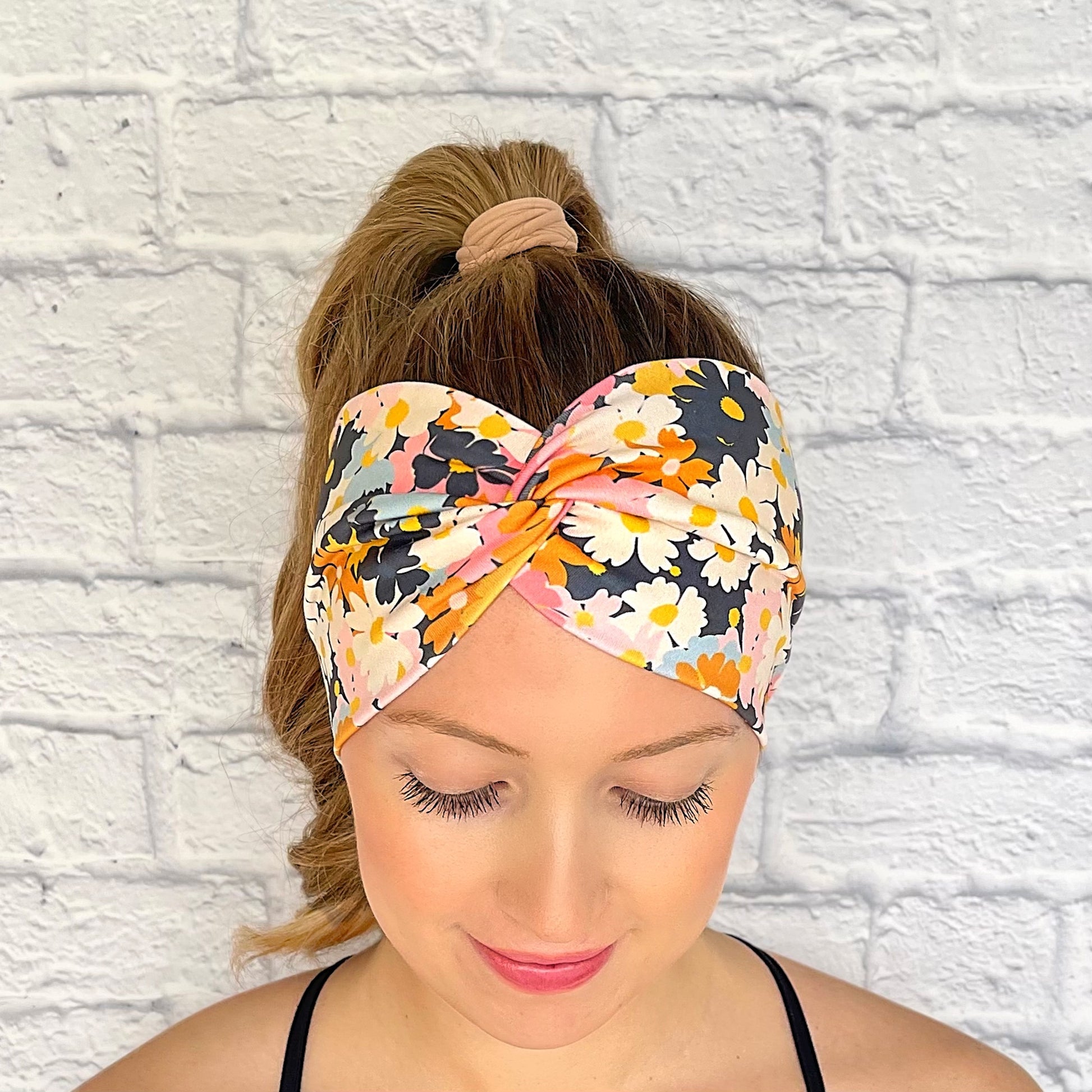 Woman with hair in curled ponytail wearing wide, twisted headband with light and dark pink, navy, light blue, white, yellow, and orange flower print.