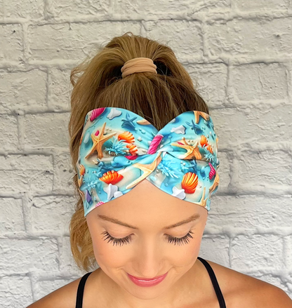 Woman with hair in ponytail wearing light blue, wide twist headband with ocean theme print- water, sand, starfish, and shells