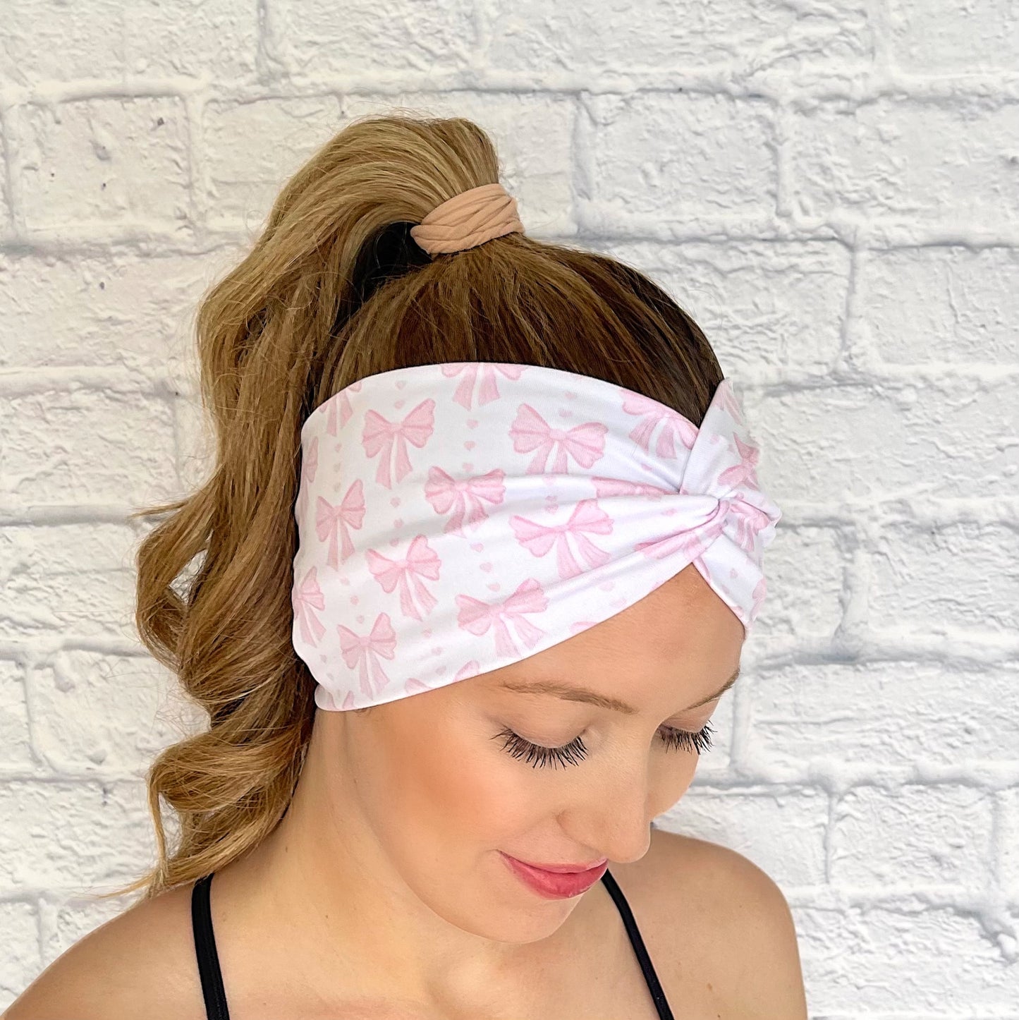 Woman with hair in curled ponytail wearing wide, twisted headband in white with light pink bow print.