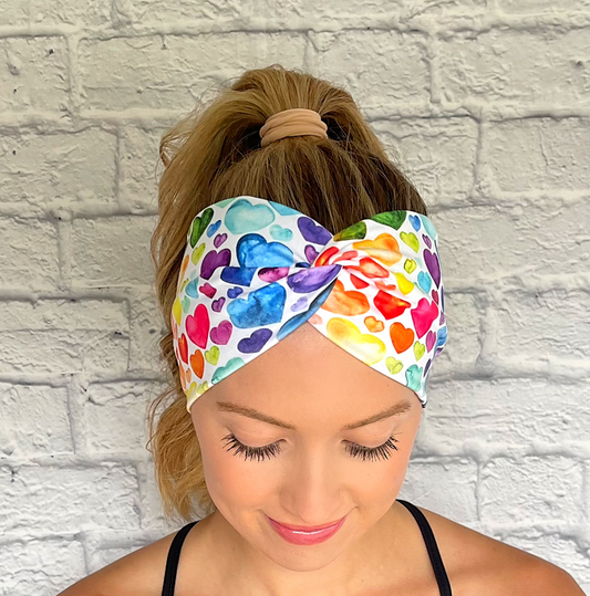 Woman with hair in curled ponytail wearing wide, twisted, white headband with rainbow heart print.