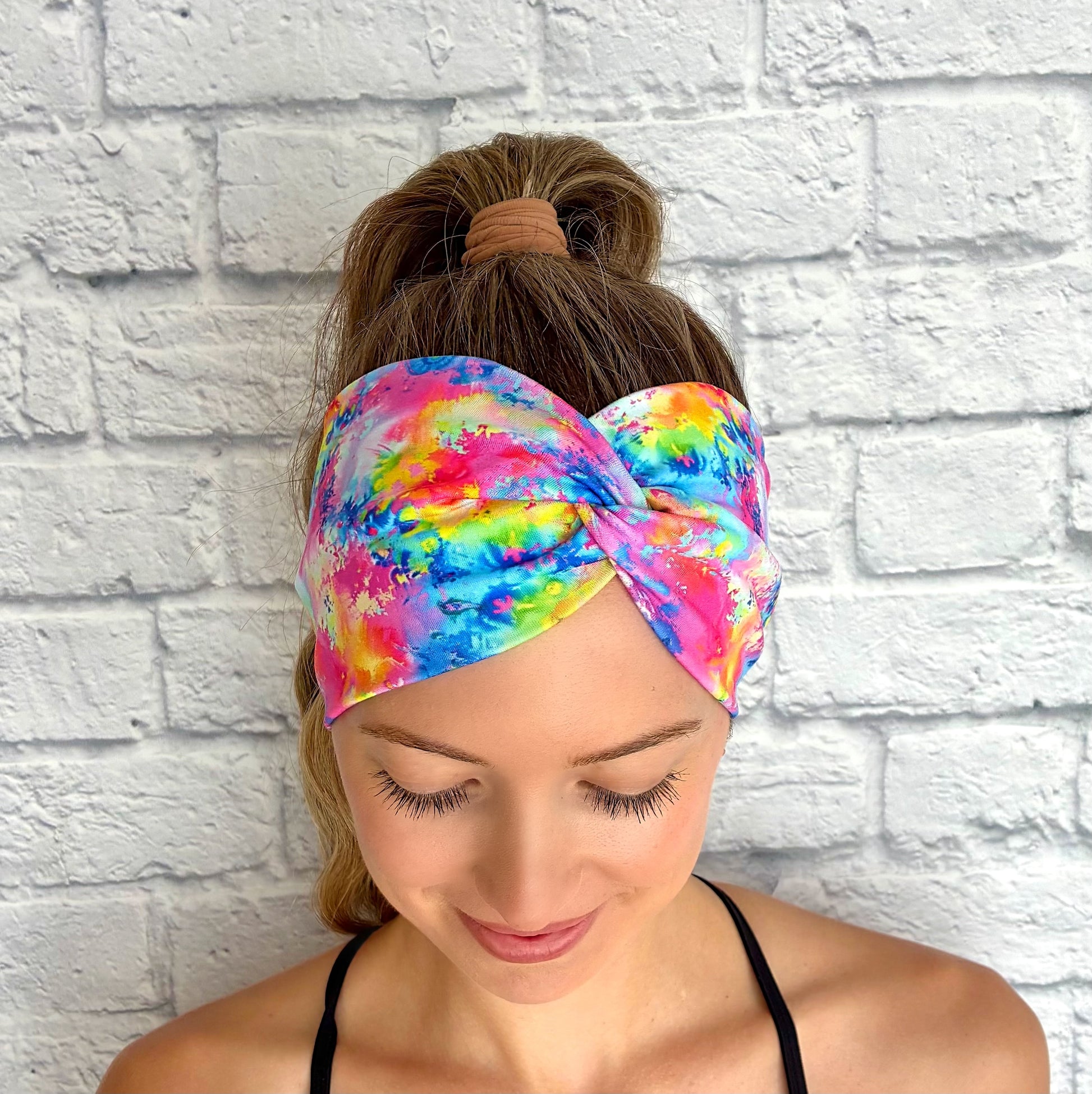 Woman wearing wide, twisted headband in rainbow color paint-splatter looking print.