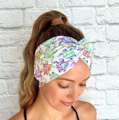 Woman with hair in ponytail wearing wide, twisted, white headband with sea turtle print, displayed on white brick background.