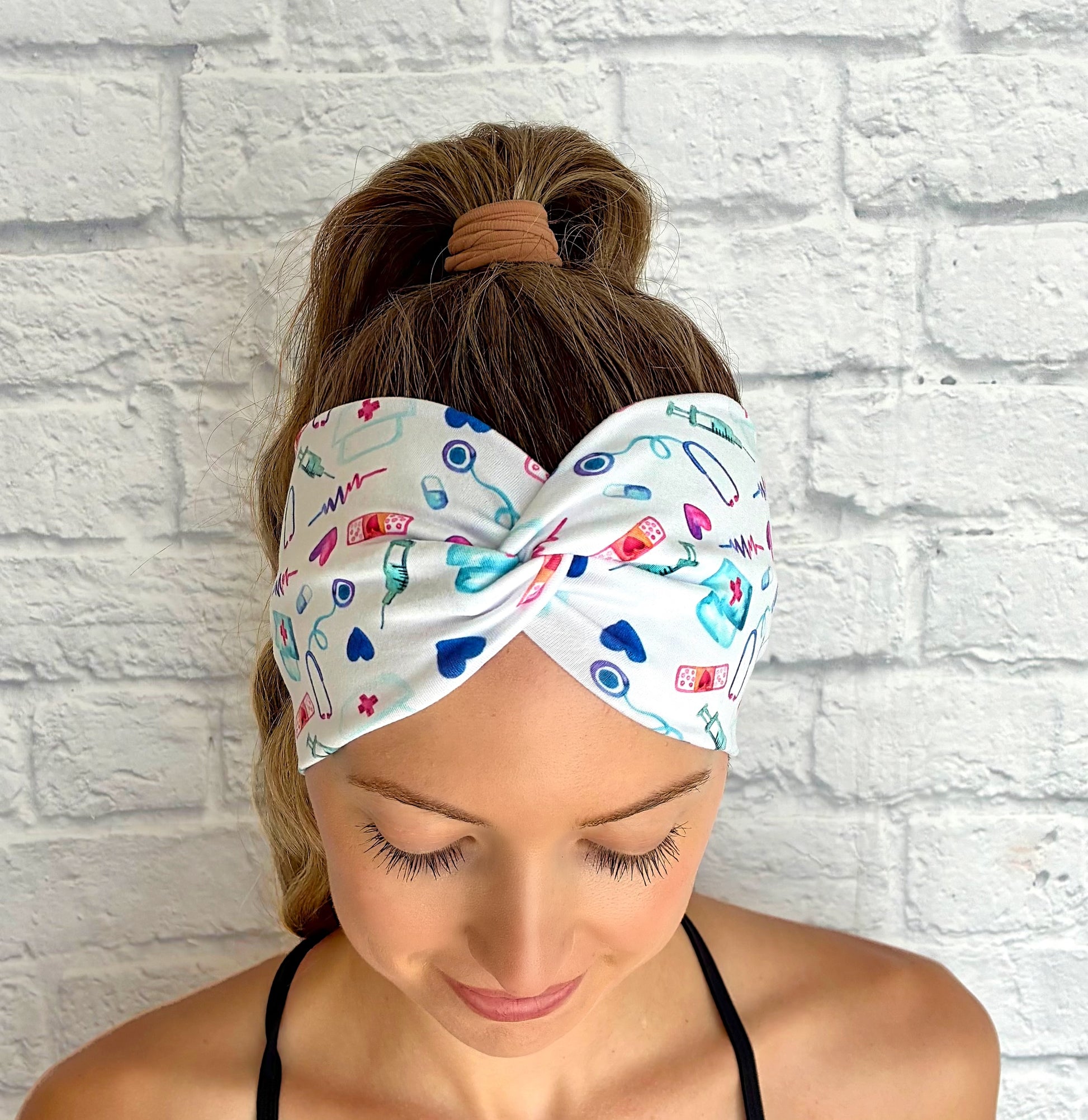 Woman with hair in ponytail wearing wide, twisted, white headband with pink, blue, and purple medical theme print.