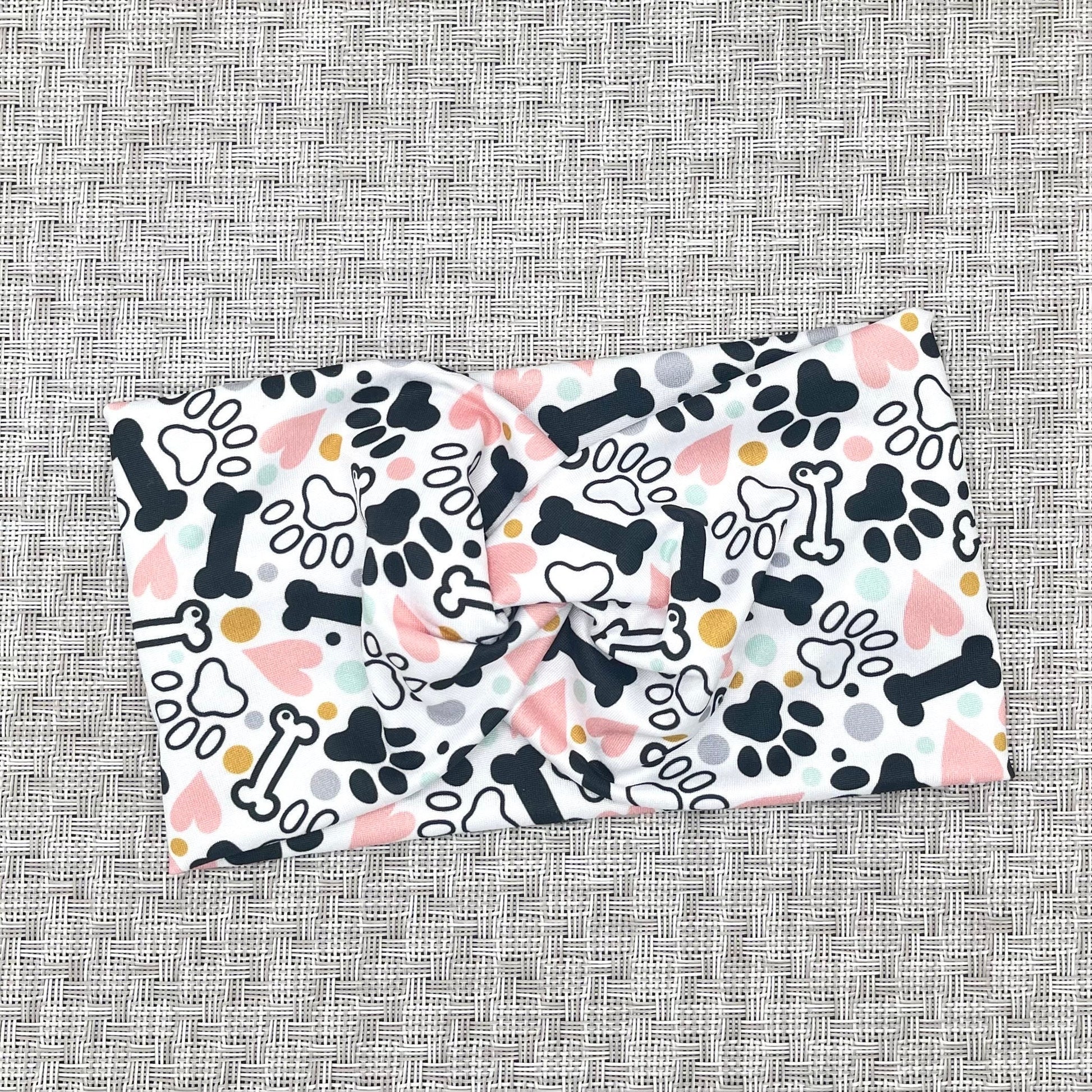 Wide, twisted headband in white with black, pink, yellow, and light blue dog theme print with paws, dog bones, and hearts.