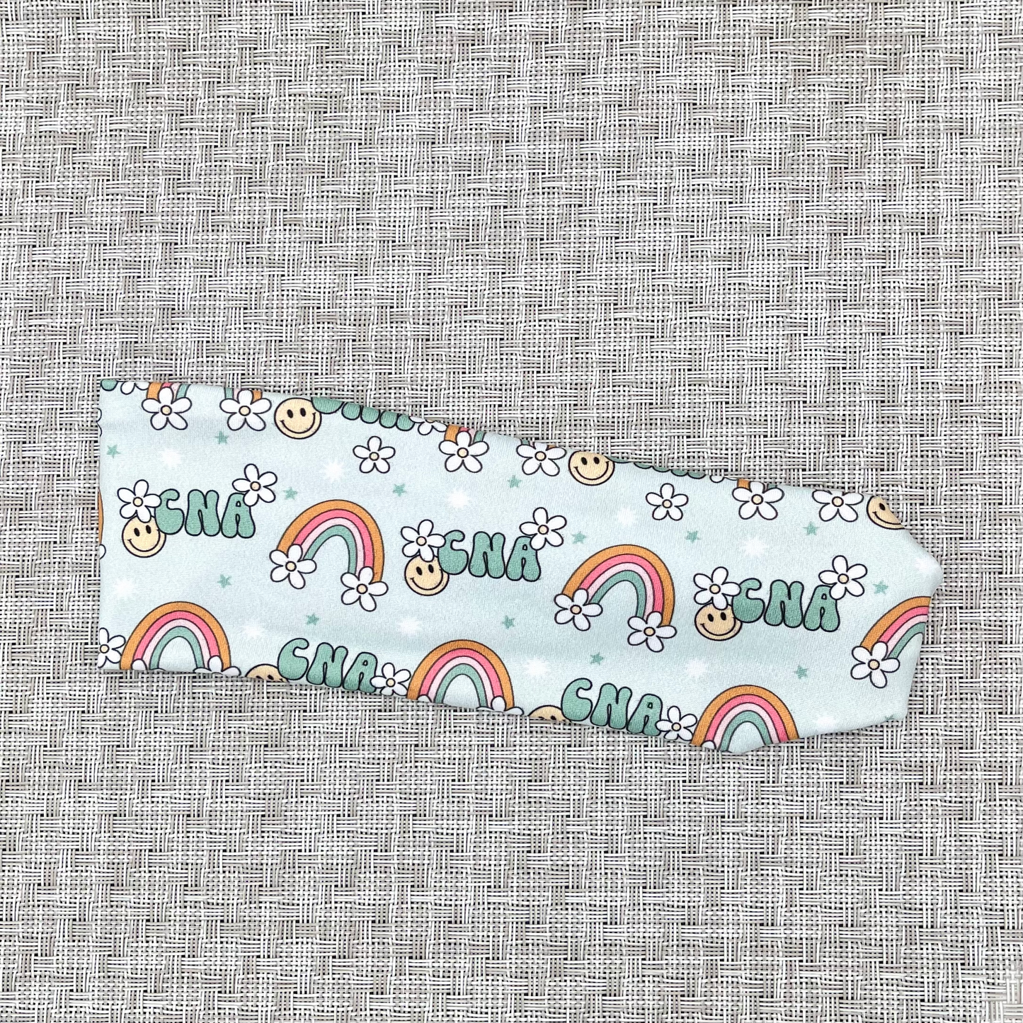 light blue headband with "cna" print, rainbows, white flowers, and smiley faces