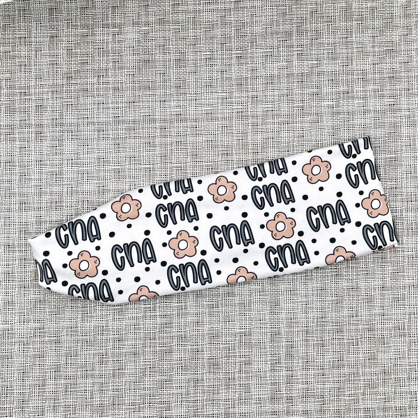 Flat, stretchy, white headband with "cna", flowers, and polka dot print in blue and tan.