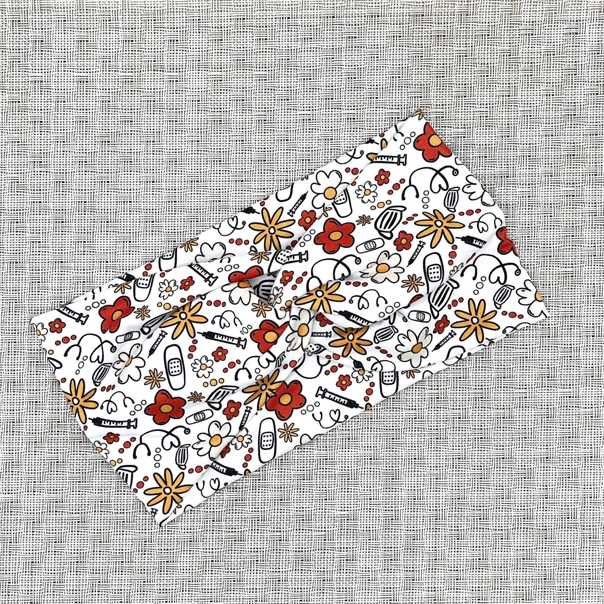 Wide, twisted headband in white with red, yellow, and black floral and medical theme print.