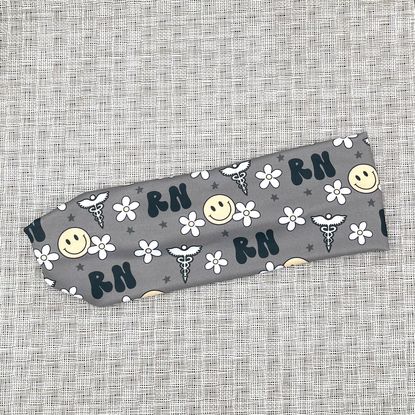 Gray, stretchy headband with black "RN" print, yellow smiley faces, white flowers, and white medical theme print.