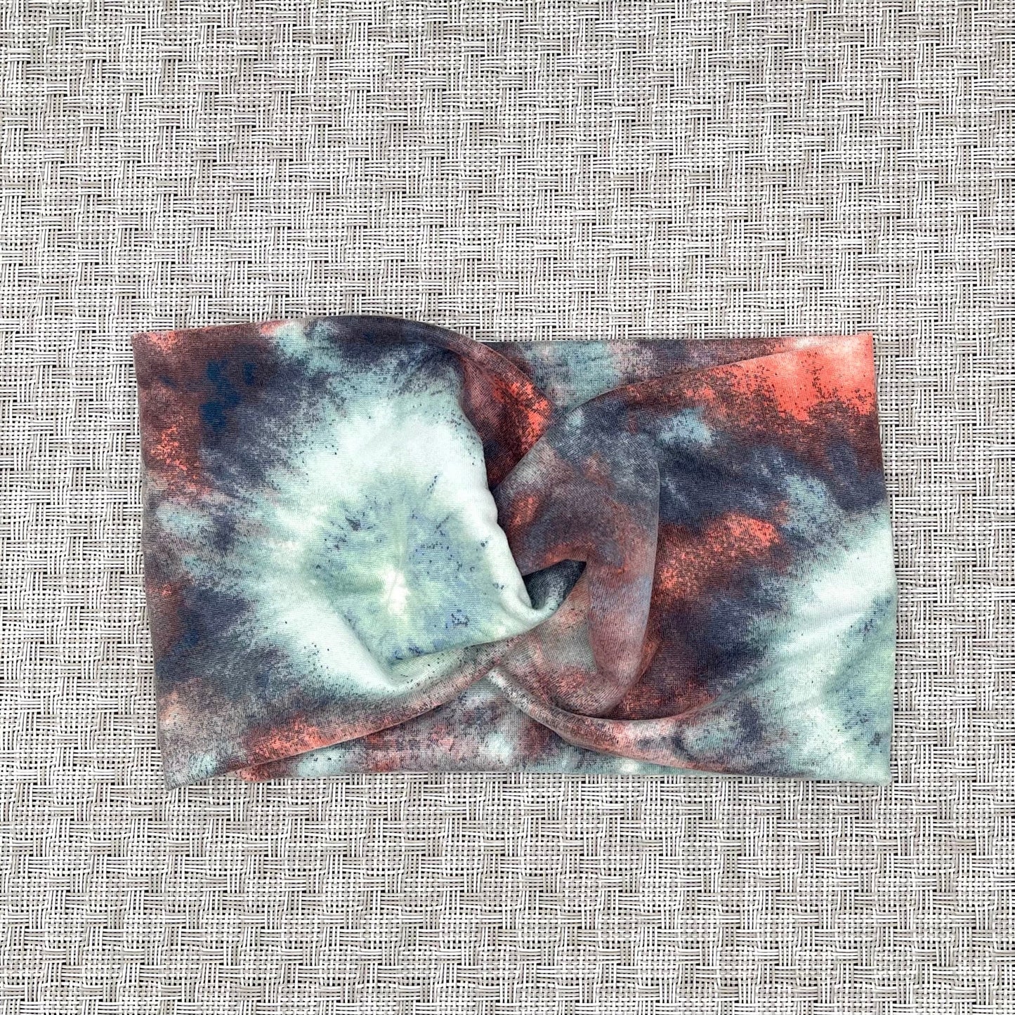 Wide, twisted headband in tie dye print with peach, white, light blue, and dark gray/blue.