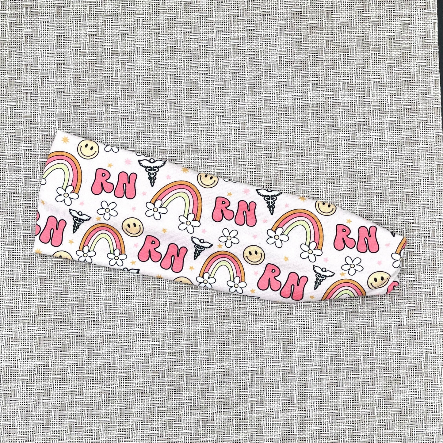 Stretchy, light pink headband with "RN", rainbows, flowers, and smiley faces