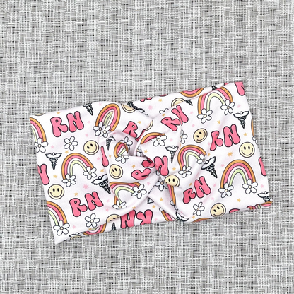 Wide, twisted headband with pink "RN" print, rainbows, flowers, smiley faces, and medical symbol.