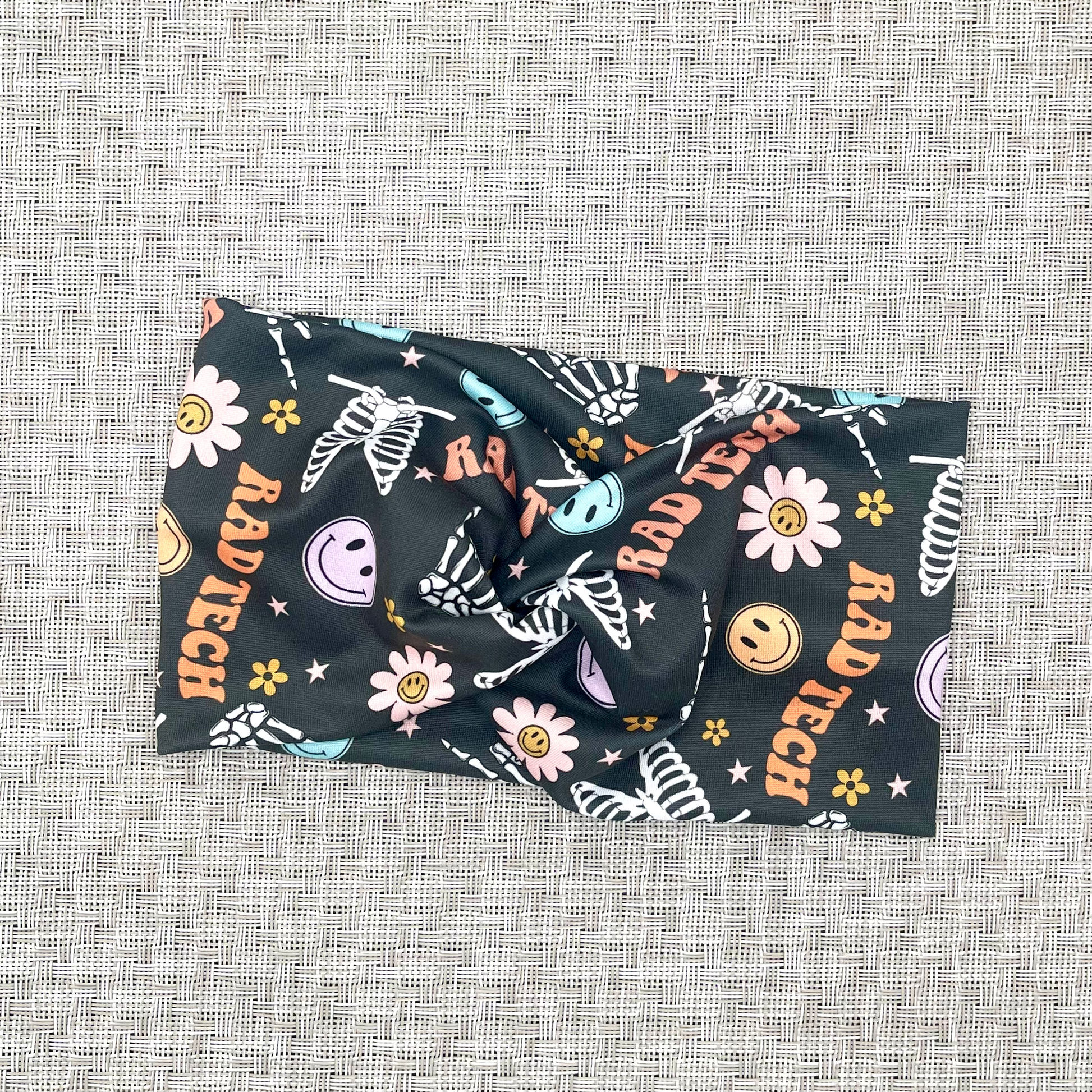 Dark gray, wide, twisted headband with skeletons, flowers, smiley faces, and "rad tech" print.