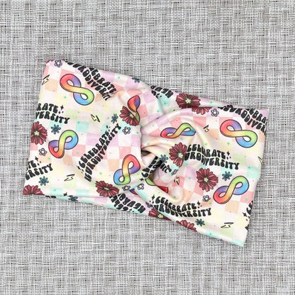 Wide, twisted headband in rainbow plaid print with "celebrate neurodiversity", rainbow infinity signs, flowers, and lightning bolt print.