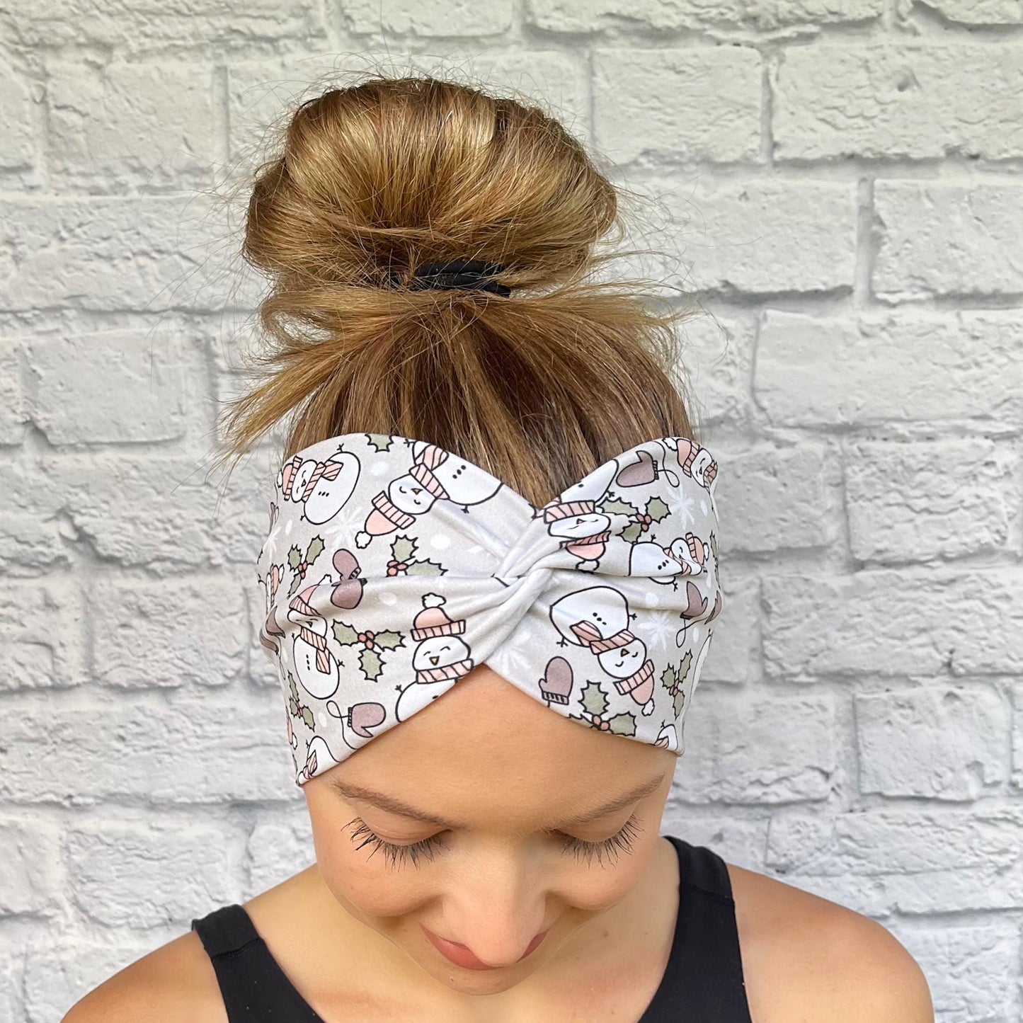 Woman with hair in bun wearing wide, twisted, gray headband with snowman print, mittens, and Christmas plants.