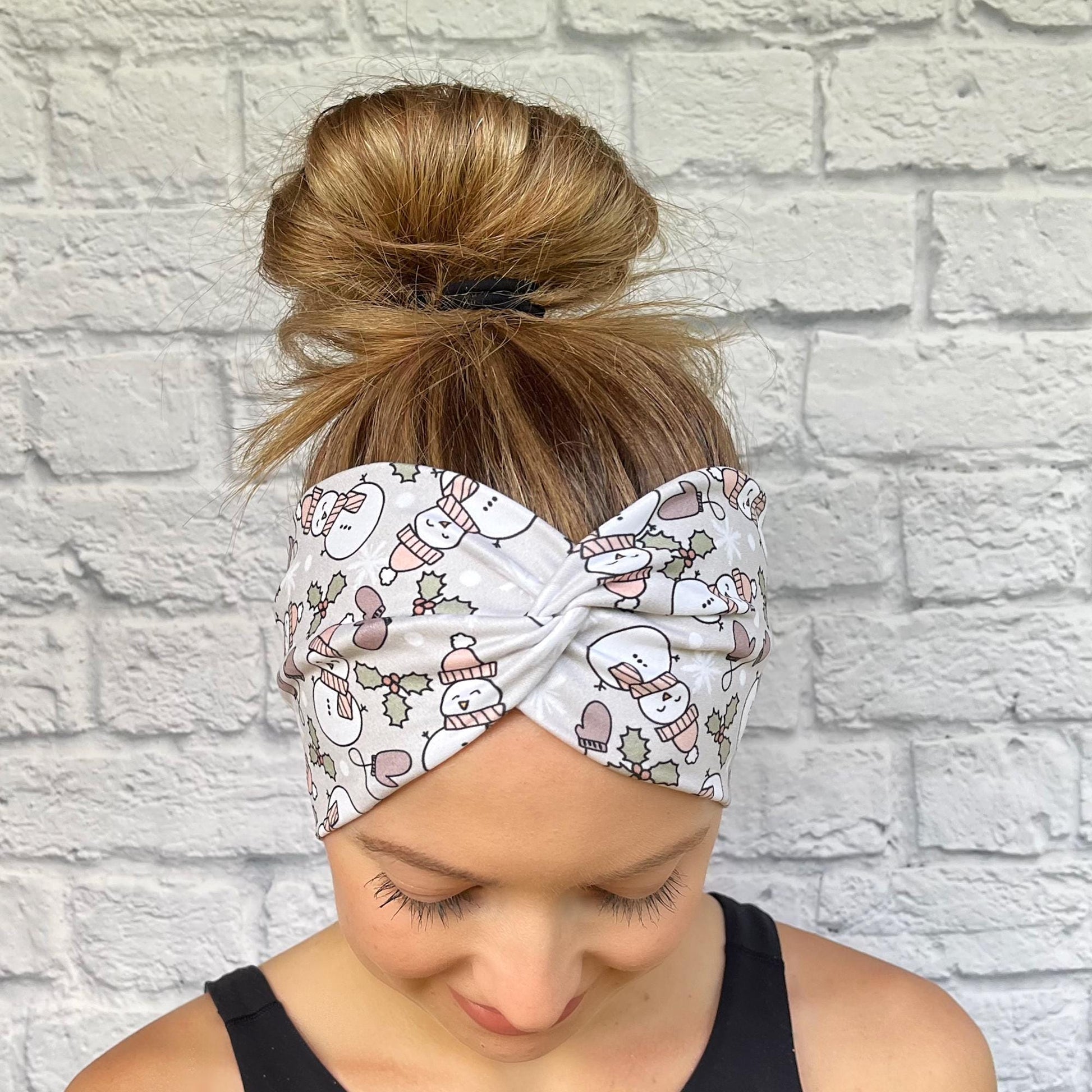 Woman with hair in bun wearing wide, twisted, gray headband with snowman print, mittens, and Christmas plants.