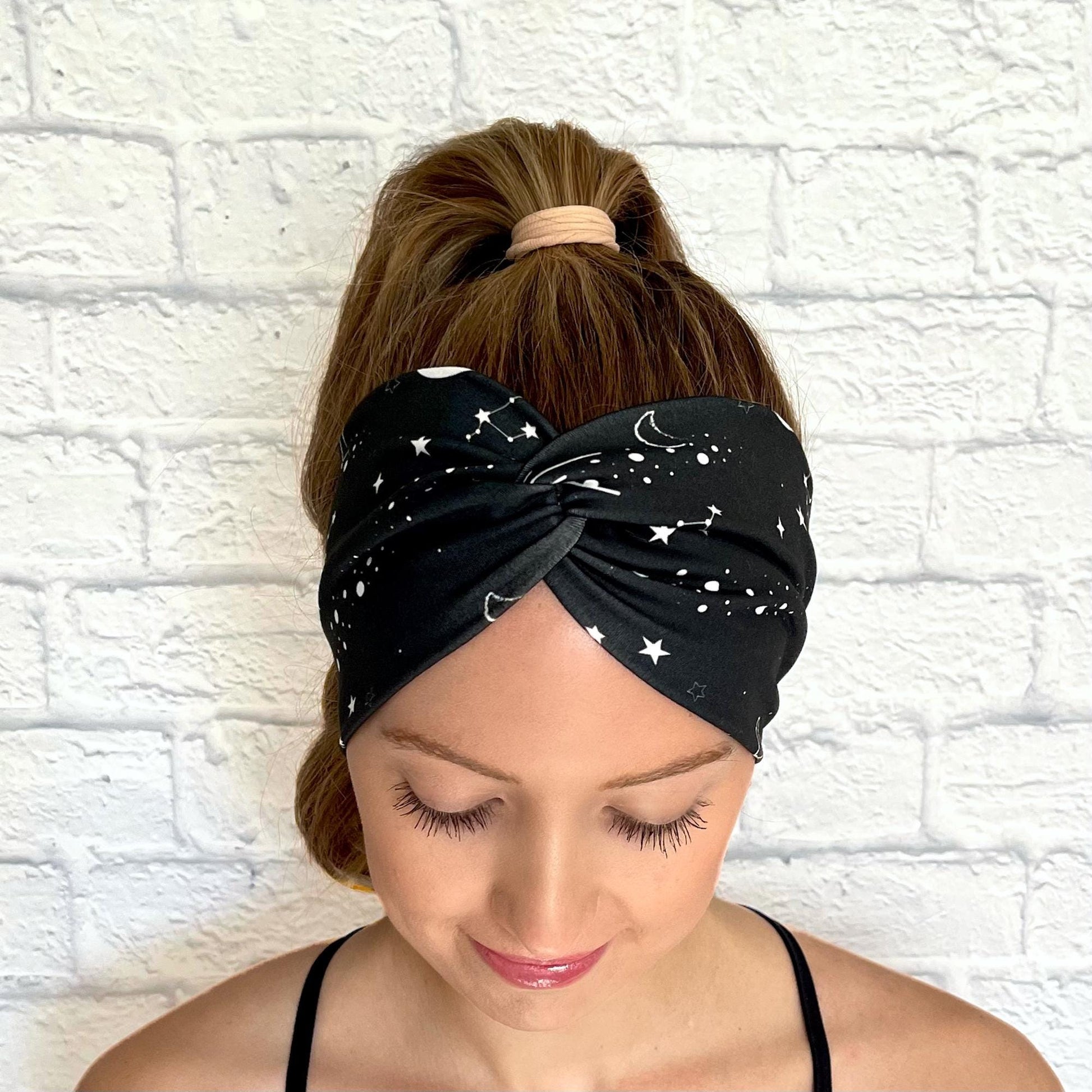 Woman with hair in curled ponytail wearing wide, twisted headband in black with white galaxy theme print.