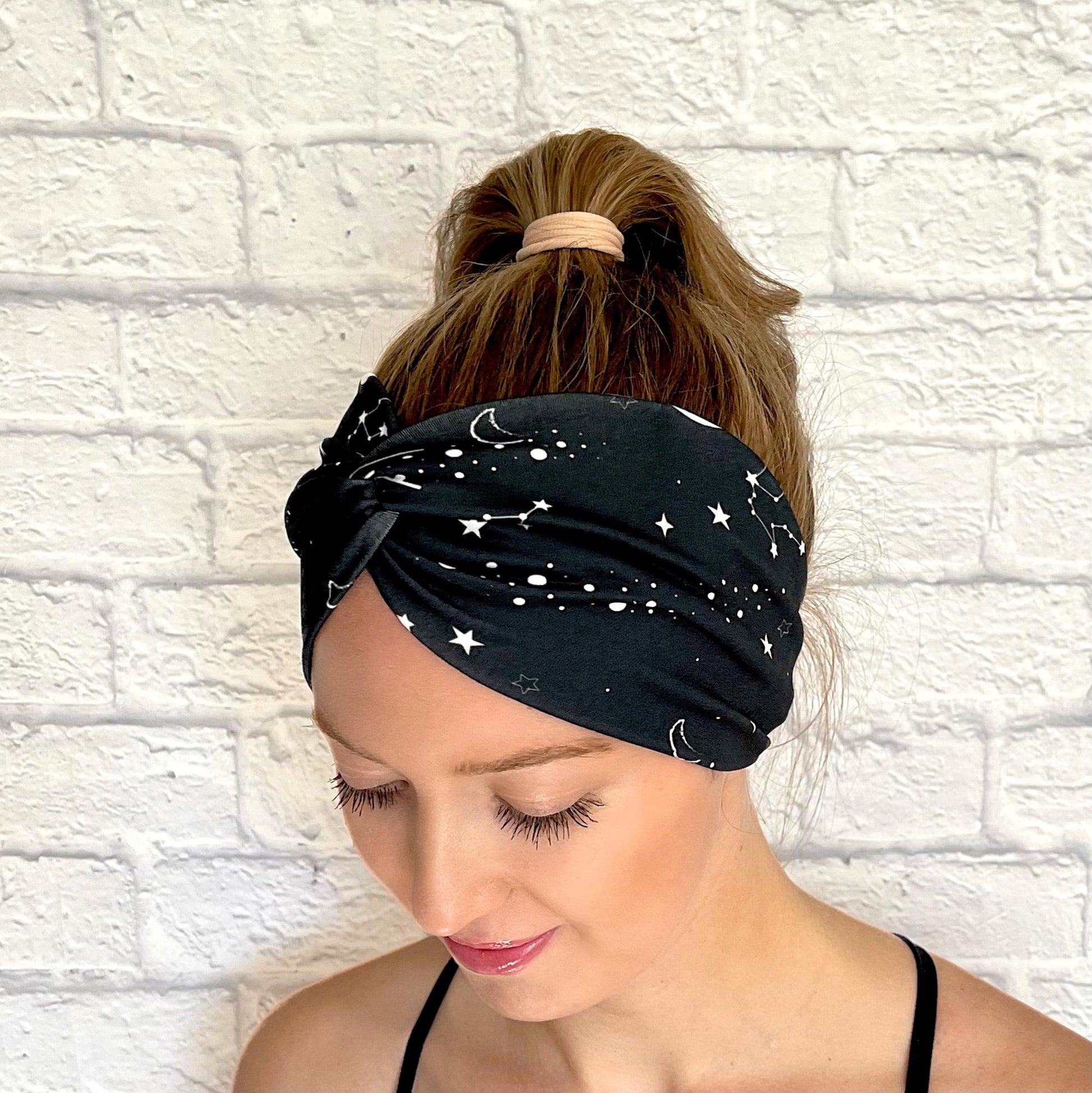 Woman with hair in curled ponytail wearing wide, twisted headband in black with white galaxy theme print.