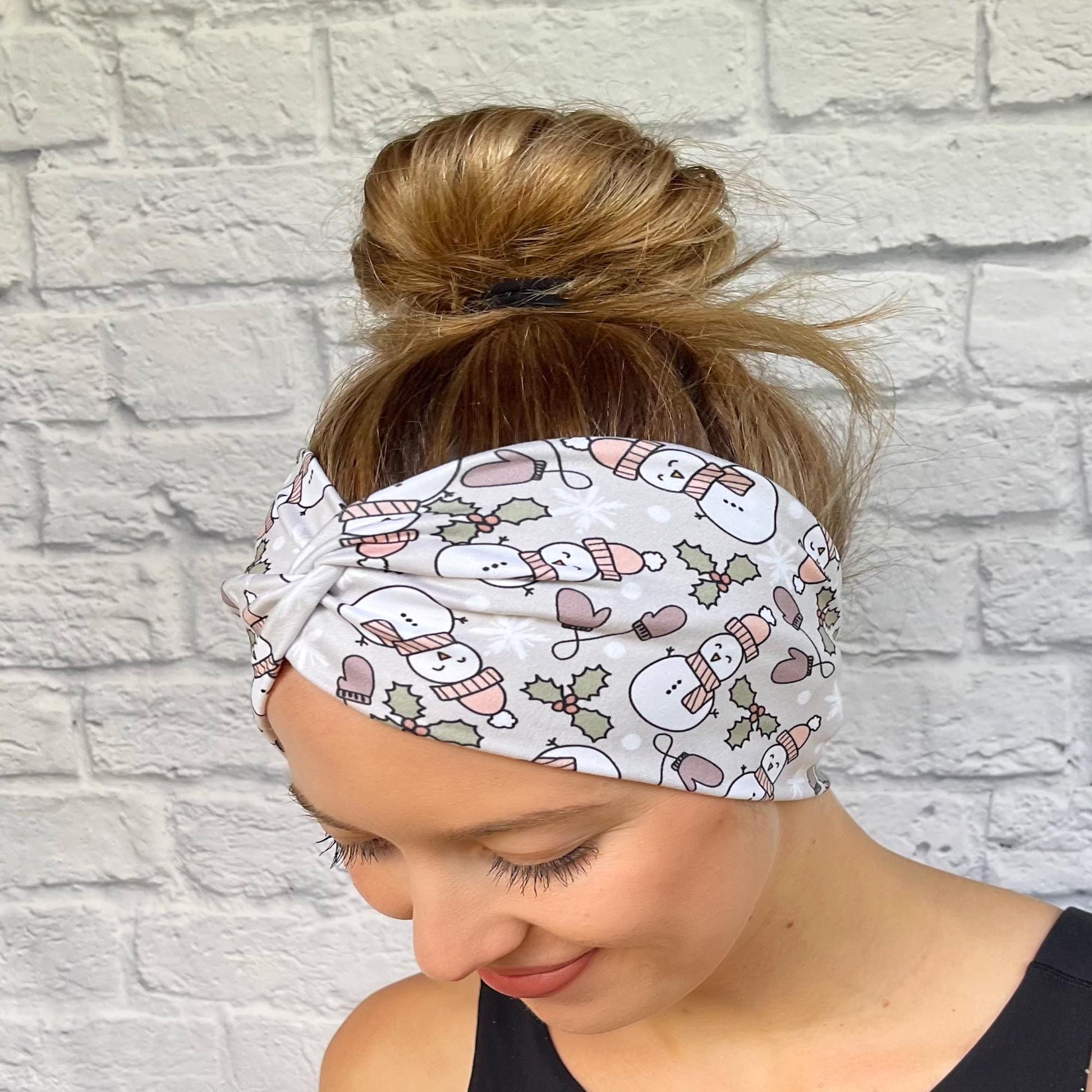 Woman with hair in bun wearing wide, twisted, gray headband with snowman print, mittens, and Christmas plants.
