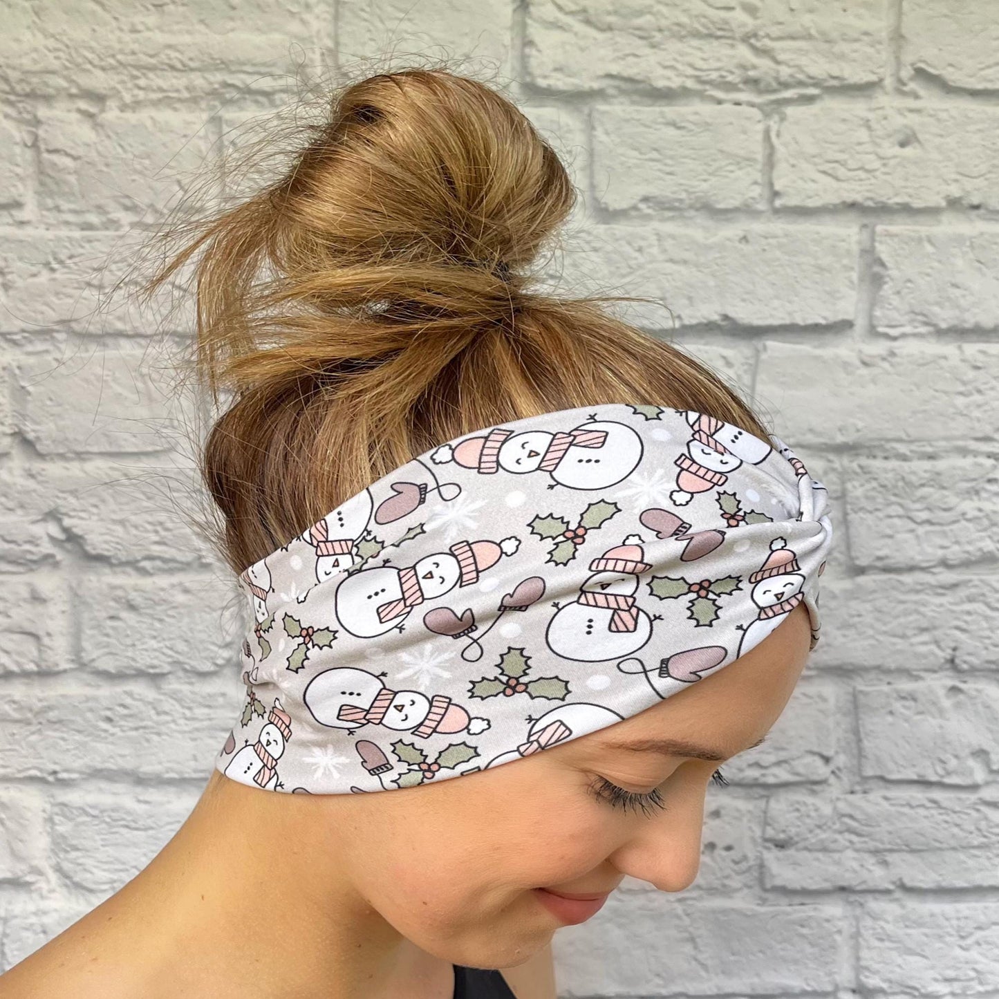 Woman with hair in bun wearing wide, twisted, gray headband with snowman print, mittens, and Christmas plants.