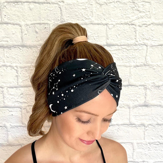Woman with hair in curled ponytail wearing wide, twisted headband in black with white galaxy theme print.