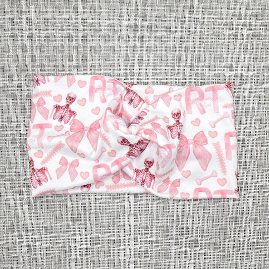 Wide, white, twisted headband with pink "RT" and "radiology tech" print, pink bows, pink hearts, and pink skeletons.