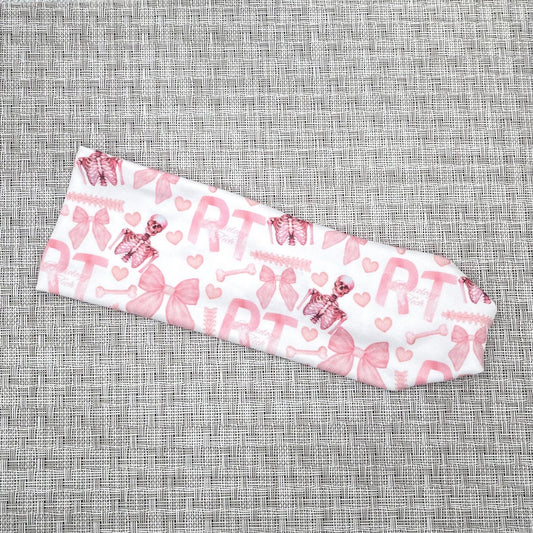 Stretchy, white headband with pink "RT" and "radiology tech" print, pink skeletons, pink bow, and pink hearts.